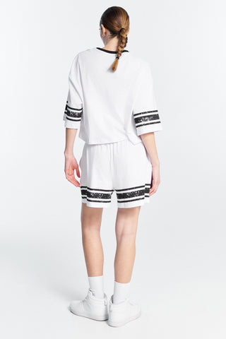 Frau trägt Lumina Sporty Shirt black, oversized mit 16 PARIS Frontprint und Kontraststreifen an Ärmeln und Saum. Kombiniert mit weißen Shorts und Schuhen für lässigen Athleisure-Look.