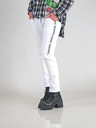 OFF#DLY Pant Raelle white: Person trägt weiße Jogpants mit elastischem Taillenbund und seitlichem Statement-Print, kombiniert mit schwarzen Stiefeln für einen modernen, sportlich-eleganten Look.