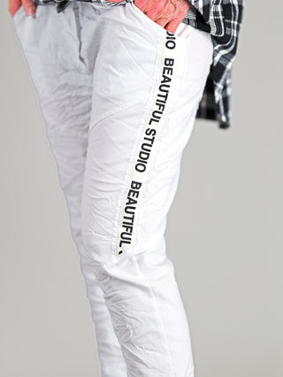 Person trägt OFF#DLY Pant Raelle white Jogpants mit elastischem Bund und seitlichem Statement-Print. Die Hose bietet eine gerade, schmale Passform mit seitlichen Eingrifftaschen, ideal für sportlich-elegante Looks.