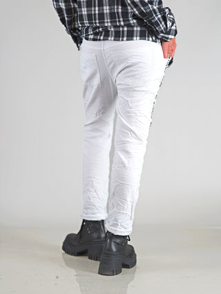 OFF#DLY Pant Raelle white: Eine Person trägt weiße Jogpants mit elastischem Bund und seitlichem Statement-Print, kombiniert mit schwarzen Stiefeln für einen sportlich-eleganten Look.