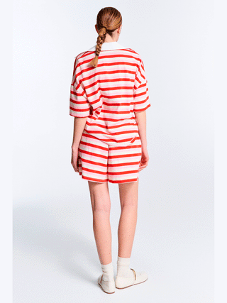 Frau trägt Lumina Polo Bluse pink/red mit roten und weißen Streifen, im modernen Oversized-Look. Sichtbar sind gestreiftes Shirt und Shorts, betont lässiger und modischer Stil.