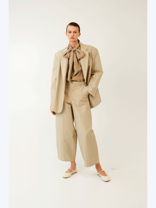 Model trägt Lumina Blazer beige, oversized geschnitten mit klassischem Reverskragen und Ein-Knopf-Schließung, aus 100% Baumwolle, kombiniert mit passender Bluse und Hose für einen minimalistischen Look.