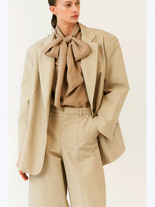 Person trägt den Lumina Blazer beige: ein hellbeiger, oversized geschnittener Blazer aus Baumwolle, kombiniert mit einer taupefarbenen Bluse und passenden, weit geschnittenen Hosen. Moderner, minimalistischer Power-Dressing-Look.