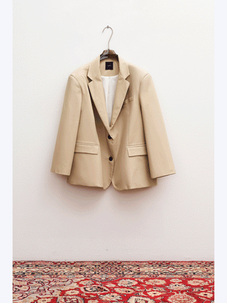 Lumina Blazer beige: Ein leichter, oversized Blazer aus 100% Baumwolle, mit klassischem Revers, zwei schwarzen Knöpfen und Pattentaschen, hängt an einem Metallbügel vor einer weißen Wand.