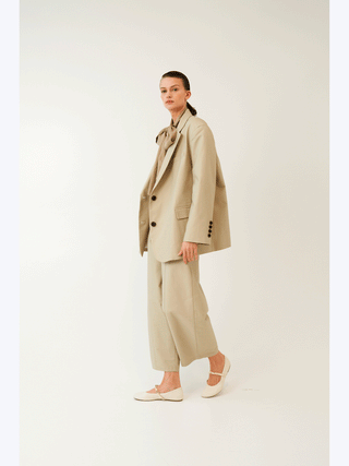 Model in übergroßem Lumina Blazer beige, unstrukturierter Schnitt mit dunklen Knöpfen, über weit geschnittenen, gekürzten Hosen. Kombiniert mit taupe Bluse und cremefarbenen Mary Jane Flats.
