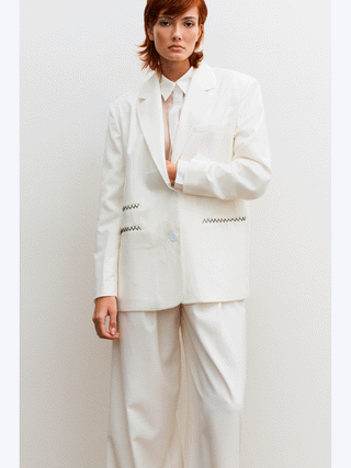 Lumina Blazer white: Person in elegantem weißen Blazer mit klassischem Reverskragen und Ein-Knopf-Schließung, dezente Kontrastnähte sichtbar, gerade Passform.
