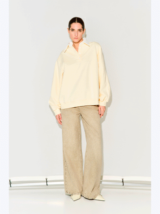 Junge Frau trägt Lumina Sweat offwhite/crema, einen oversized Baumwoll-Polosweater mit weißem Kragen und Knopfleiste, kombiniert mit weiten, steinfarbenen Jeans und spitzen Schuhen, vor hellem Hintergrund.