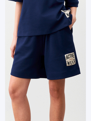 Person trägt Lumina Activ Short blue mit „Active Wear“-Patch. Shorts haben einen bequemen, weiten Schnitt und elastischen Bund, ideal für sportliche und freizeitliche Aktivitäten.
