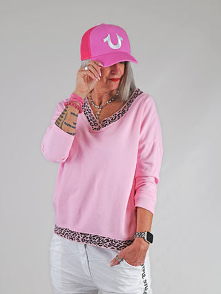Frau trägt OFF#DLY Lightsweat Liora rose/leo, ein locker geschnittenes Sweatshirt mit Leo-Details an Ausschnitt und Saum, kombiniert mit einer rosa Mütze. Ideal für lässigen Chic und Komfort.