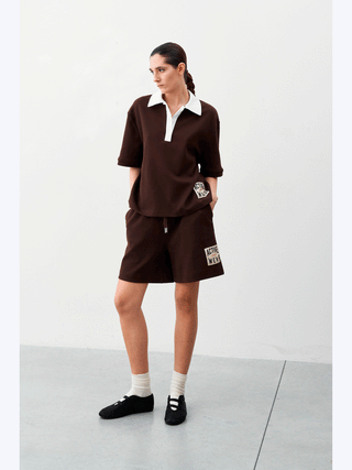 Frau in braunem Lumina Activ Short, sportlicher Schnitt mit elastischem Bund, trägt schwarze Schuhe und weiße Socken. Sichtbar ist auch ein „Active Wear“-Patch als urbanes Design-Detail.