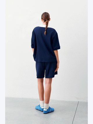Frau trägt Lumina Activ Short brown/white mit elastischem Bund und sportlichem Schnitt, kombiniert mit blauer Bluse und weißen Socken. Ideal für sportliche Freizeitaktivitäten.