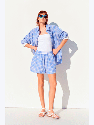 Frau trägt die Lumina Bluse checker blue, ein kariertes Oversized-Hemd mit umkrempelbaren Manschetten, kombiniert mit Shorts und Sandalen. Ideal für lässigen Sommerstil.