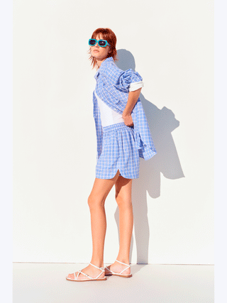 Frau trägt Lumina Bluse checker blue, ein oversized kariertes Hemd mit Knopfleiste und umkrempelbaren Manschetten. Kombiniert mit lässigem Look, perfekt für sommerliche Outfits.