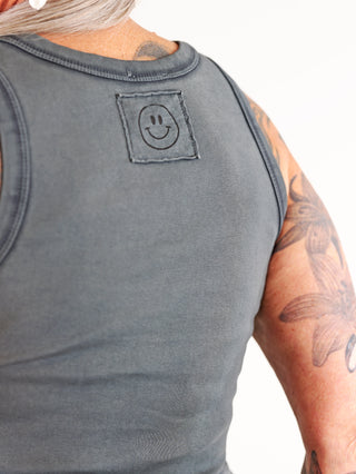 Eine Person trägt das OFF#DLY Top Neve wash black, ein graues Baumwoll-Tanktop mit aufgenähtem Smiley-Patch auf dem Rücken und einem lässigen Rundhalsausschnitt.