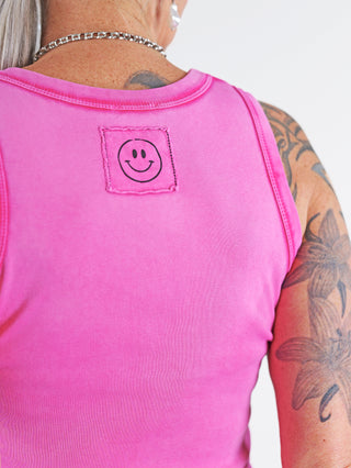 Person trägt das OFF#DLY Top Neve pink, ein ärmelloses Baumwoll-Top mit aufgenähtem Smiley-Patch. Sichtbare Tattoos und eine Silberkette ergänzen den lässigen Look.