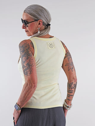 Frau mit grauen Haaren trägt das OFF#DLY Top Neve gelb, ein lässiges Baumwoll-Top mit Smiley-Patch. Tattoos am Arm und modisches Accessoire betonen den Look.