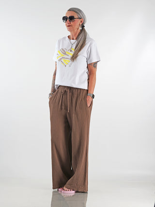 Frau trägt die OFF#DLY Musselin Pant Frida mokka, eine luftige Wide-Leg-Hose aus Baumwolle mit elastischem Bund und Kordelzug, ideal für entspannte und stilvolle Outfits.