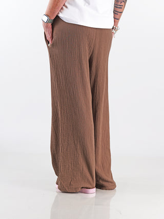 Person trägt OFF#DLY Musselin Pant Frida mokka, eine luftige Wide-Leg-Hose mit elastischem Bund und Seitentaschen, kombiniert mit weißem Hemd und Uhr, perfekter Stil für warme Tage.