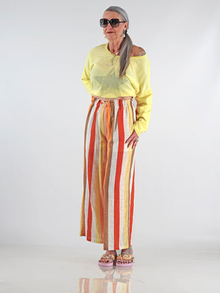 Frau trägt die OFF#DLY Leinenhose Duffy multicolor mit weiten, farbenfrohen Streifen, kombiniert mit Sandalen. Die Hose hat einen elastischen Bund und eine integrierte Innenhose für Komfort.
