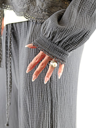Nahaufnahme einer Hand mit langem Nagel, einem Perlenring und einem grauen Musselin-Ärmel. Zeigt die OFF#DLY Musselin Jacke Ritta anthrazit mit dekorativer Spitzenborte und weiten Ärmeln.
