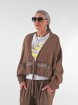 Frau trägt die OFF#DLY Musselin Jacke Ritta mokka mit weiter Silhouette, Spitzenborte und Häkel-Details, kombiniert mit Sonnenbrille und braunem Oberteil. Jacke verleiht femininen, luftigen Look.