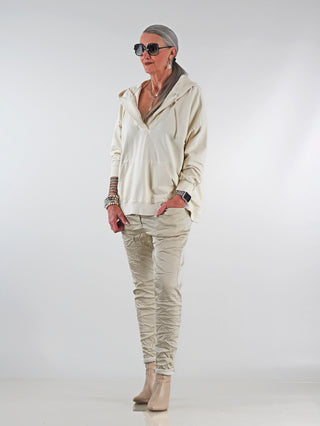 Frau in cremefarbenem OFF#DLY Sweat Ines Hoodie mit Oversize-Schnitt, Kapuze und Kängurutasche, kombiniert mit gestreiften Hosen, trägt Armbanduhr und Sonnenbrille. Stilvoll und lässig.