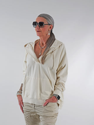 Frau trägt OFF#DLY Sweat Ines creme Hoodie mit Kapuze und Kängurutasche, kombiniert mit Sonnenbrille und Schmuck, präsentiert lässigen Oversize-Look.
