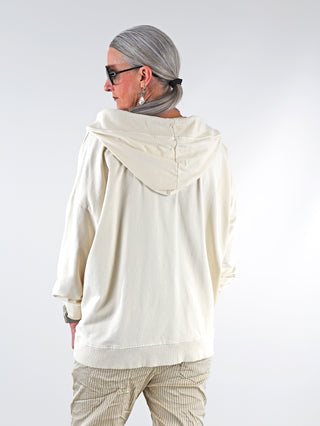 Frau trägt OFF#DLY Sweat Ines creme, oversized Hoodie mit Kapuze und Kängurutasche, ideal für lässigen Stil. Kombiniert mit Accessoires und Boots für eleganten Look.