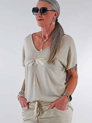 Frau trägt das OFF#DLY Shirt Libella beige mit glitzernder Libellenapplikation und V-Ausschnitt. Kombiniert mit Sonnenbrille und Schmuck, präsentiert es einen lässig-eleganten Stil.