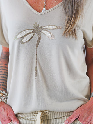 Person trägt das OFF#DLY Shirt Libella beige mit V-Ausschnitt und glitzernder Libellenapplikation, ideal für lässige Looks. Kombiniert mit einer Perlenkette für einen eleganten Touch.