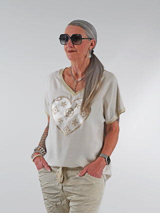 OFF#DLY Shirt Heart beige: Frau trägt ein T-Shirt mit Herz-Print und Goldglitzer-Details, kombiniert mit einer Perlenkette, Sonnenbrille und Armbändern. Ideal für elegante Alltagslooks.