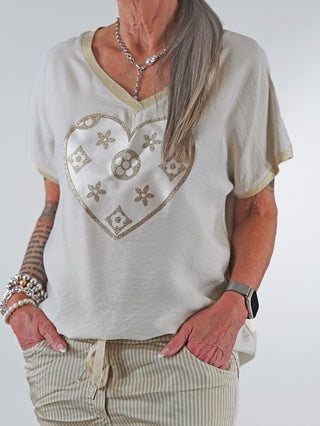 Frau trägt OFF#DLY Shirt Heart beige mit großem Herz-Print und Glitzer, V-Ausschnitt mit Lurexkante, leicht überschnittene Schultern, Arme in den Taschen. Perfekt für eleganten, femininen Look.