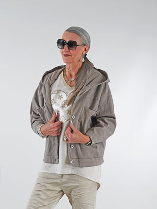 Frau trägt die OFF#DLY Jacke Zuleika taupe mit Kapuze, großen Taschen und silbernen Druckknöpfen. Die Jacke hat einen lässigen, kastigen Schnitt, der bis Größe 44 passt.