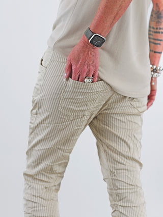 OFF#DLY Pant Stripes Gitta beige: Person trägt gestreifte, beige Hose mit elastischem Bund und schmal zulaufendem Bein, Hand in der Hosentasche, Uhr am Handgelenk sichtbar.