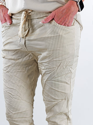 OFF#DLY Pant Stripes Gitta beige: Person steckt Hände in beige Streifenhose mit elastischem Bund und schmal zulaufendem Bein. Details wie Seitentaschen und Kordelzug erkennbar.