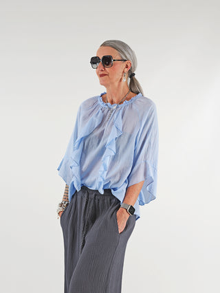 Frau trägt die OFF#DLY Rüschen Bluse Frida lightblue aus fließender Viskose mit Volants und 3/4-Ärmeln, kombiniert mit Sonnenbrille, sichtbar in entspannter Silhouette.