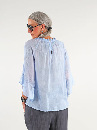 Frau trägt die OFF#DLY Rüschen Bluse Frida lightblue mit Volants und weiten 3/4-Ärmeln, kombiniert mit Sonnenbrille und Ohrringen, perfekt für sommerliche Eleganz.