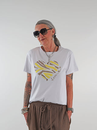 Frau trägt das OFF#DLY Tee Heart white mit glitzerndem Herzmotiv, kombiniert mit braunen Hosen. Sie hat graue Haare und trägt große Sonnenbrillen, eine Halskette und Armbänder.