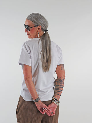 Frau mit langen Haaren trägt eine Sonnenbrille und ein weißes OFF#DLY Tee Heart. Armbänder betonen das Outfit.