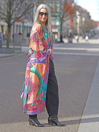 Frau trägt OFF#DLY Kimono Adelina multicolor im Boho-Stil mit tropischem Print, weiten Ärmeln und fließender Viskose. Kombiniert mit schwarzen Stiefeln und Sonnenbrille, draußen fotografiert.