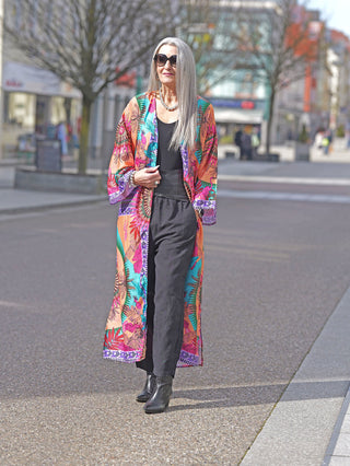 Frau trägt OFF#DLY Kimono Adelina multicolor in Boho-Stil, mit weitem Schnitt und exotischem Muster, ergänzt durch schwarze Hosen und Sonnenbrille, während sie draußen auf der Straße steht.