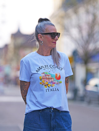 Frau trägt OFF#DLY Tee Amalfi white mit Amalfi-Print, der Zitrone und Aperol zeigt. Sie trägt Sonnenbrille und weiße Bluse, mit Jeans kombiniert. Ideal für Italien-Fans.