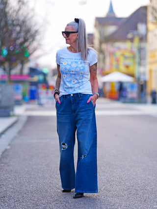 Frau auf Straße trägt das OFF#DLY Flower Market white/blue T-Shirt mit floralen Print und Glanzeffekt, kombiniert mit blauen Jeans. Ideal für entspannte Frühlingsmode.