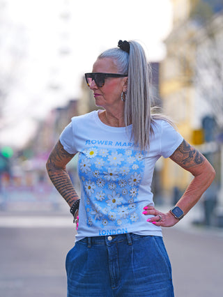 OFF#DLY Flower Market white/blue T-Shirt mit Blumenprint, getragen von tätowierter Frau mit Sonnenbrille. Kombiniert mit blauen Jeans, vermittelt es einen frischen, frühlingshaften Look.
