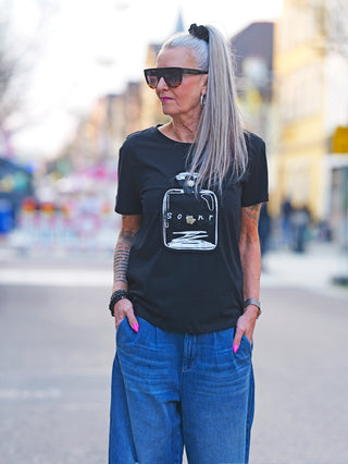 Frau trägt das OFF#DLY Tee Flacon black mit Flacon-Print, verziert mit Satinblume und Schmetterlingsbrosche. Kombiniert mit Sonnenbrille und schwarzem Oberteil, Hände in den Taschen.