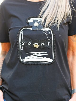 Frau trägt OFF#DLY Tee Flacon black mit weiß-silbernem Flacon-Print, Satinblume und abnehmbarer Schmetterlingsbrosche. Ideal für stilvolle Casual-Looks, Einheitsgröße bis Größe 40.
