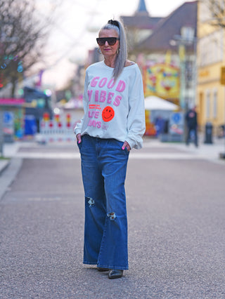 Frau trägt OFF#DLY Sweat Good Vibes white: weißer Oversize-Sweater mit rosa GOOD VIBES Schriftzug und orangefarbenem Smiley, kombiniert mit blauer Jeans, modisch und lässig.