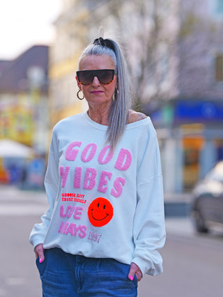 Frau trägt OFF#DLY Sweat Good Vibes white, einen weißen Oversize-Sweater mit GOOD VIBES und Smiley in 3D-Frottee. Kombiniert mit Jeans für lässigen Stil.