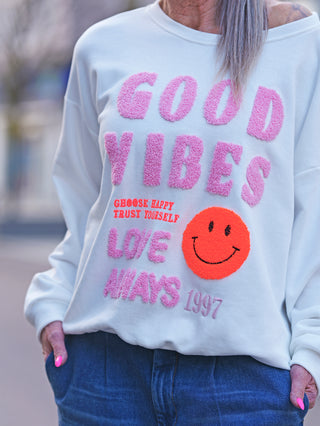 Person trägt OFF#DLY Sweat Good Vibes white, einen cremeweißen Oversize-Sweater mit rosa GOOD VIBES-Schriftzug und fröhlichem Smiley-Patch, kombiniert mit blauen Jeans.
