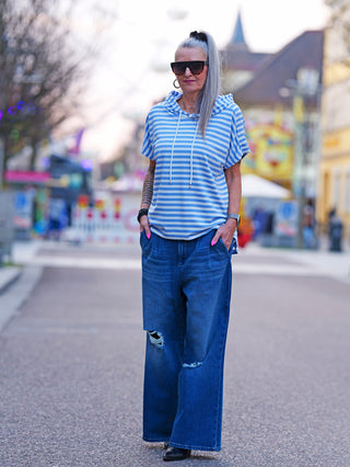 Frau auf der Straße trägt das OFF#DLY Ringel m. Kapuze white/lightblue, ein gestreiftes Kurzarm-Shirt mit Kapuze, kombiniert mit blauen Jeans und Sonnenbrille. Ideal für einen lässigen, maritimen Look.